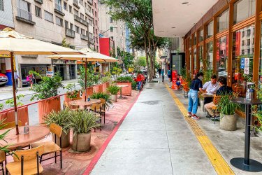 imagem de calçada. ao lado esquerdo mesas e plantas ao meio caminho de passagem, mais a direita sinalização de acessibilidade e mais a direita o estabelecimentos com algumas mesas fora