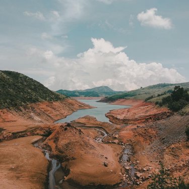 imagem montanhas, ao meio corre um rio sinuoso e ao fundo da imagem ele aumenta e mais ao fundo mostra outras montanhas ao longe. Além do céu encoberto por nuvens brancas.